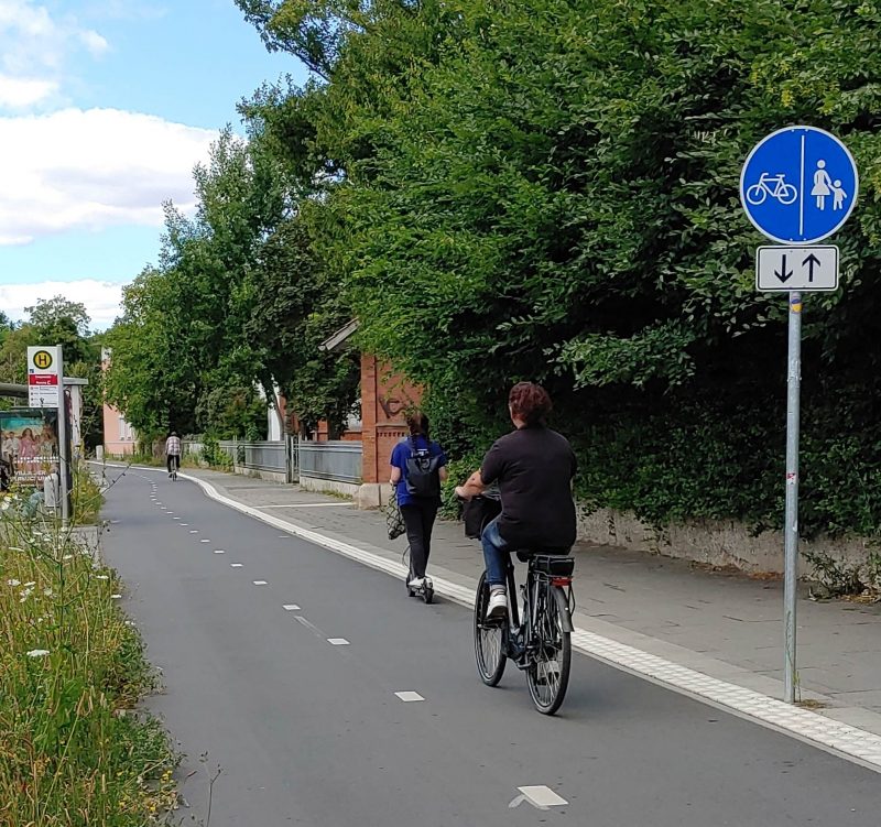2 Radfahrende und eine Person auf einem E-Scooter fahren auf einem breiten und gut ausgebautem Radweg. Der Radweg ist durch einen Mittelstreifen gekennzeichnet und in beide Richtungen befahrbar.